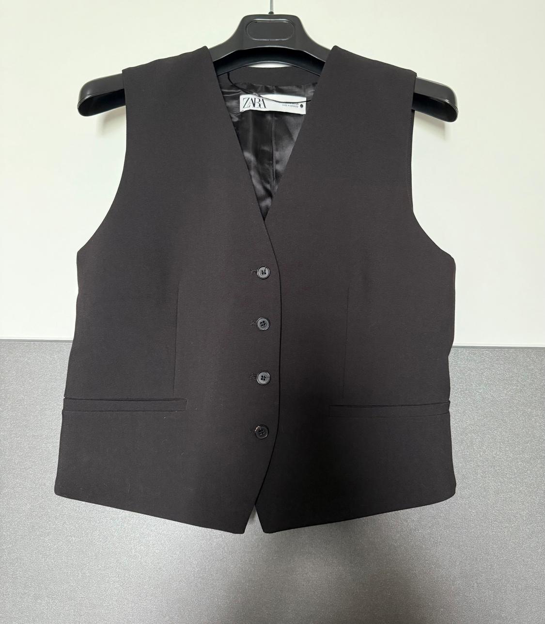 Black sleeveless waistcoat with five buttons and front pockets, hanging on a black hanger. Visible label with text "ZARA".