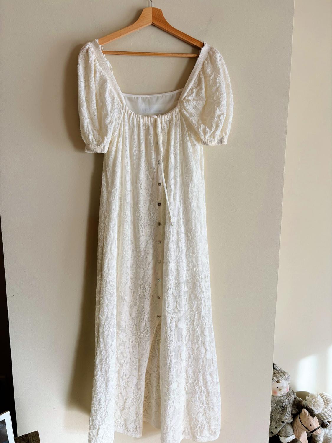 White lace dress with puff sleeves and front buttons, hanging on a wooden hanger against a beige wall.