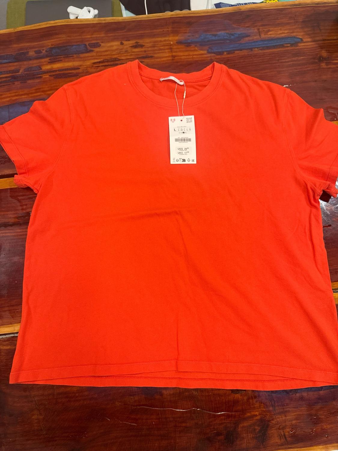 Vibrant orange short-sleeved basic t-shirt, laid out flat on a wooden table. Label with text: L US$ 29.90 UK£ 17.99.