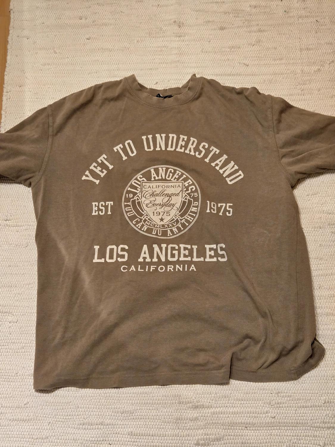 Καφέ μπλουζάκι με λευκό κείμενο: "Yet to Understand", "Los Angeles California", "Challenged Everyday", "Est 1975", πάνω σε μπεζ χαλί.