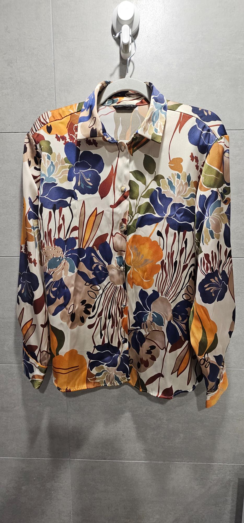 Long-sleeved shirt with a floral print in shades of blue, orange and green, hanging on a hanger against a grey tiled wall.