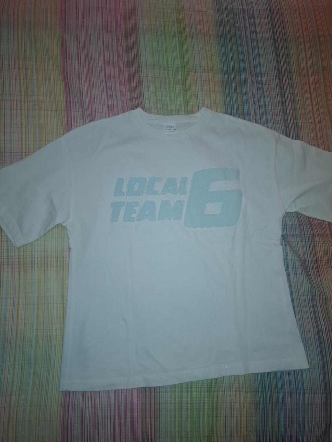 White short-sleeved T-shirt with the text ‘LOCAL TEAM 6’ in light blue, against a fabric background with coloured stripes.