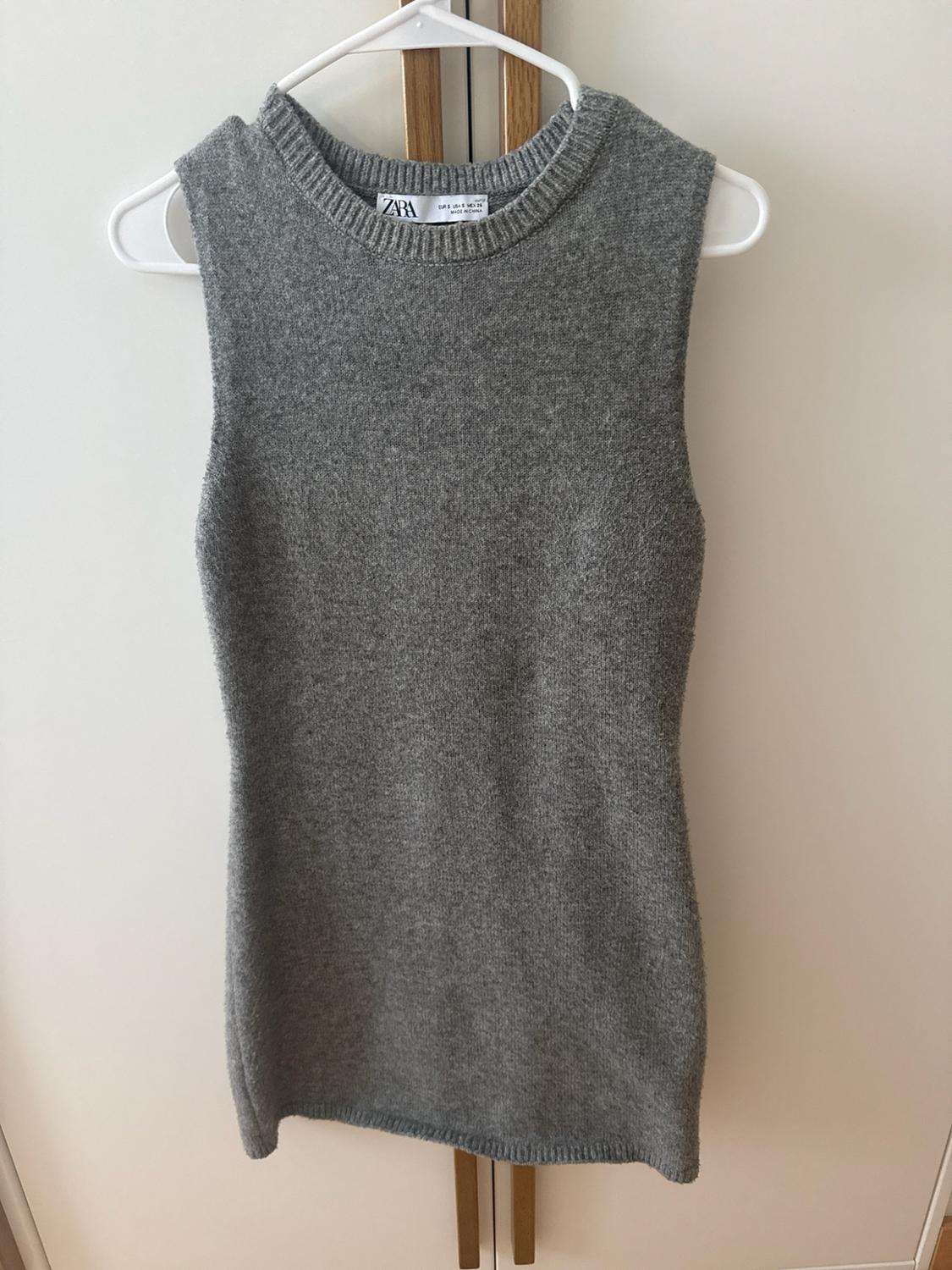 Grey sleeveless knitted top with a round neck hanging on a white hanger in front of a wardrobe. Label with the text ZARA.