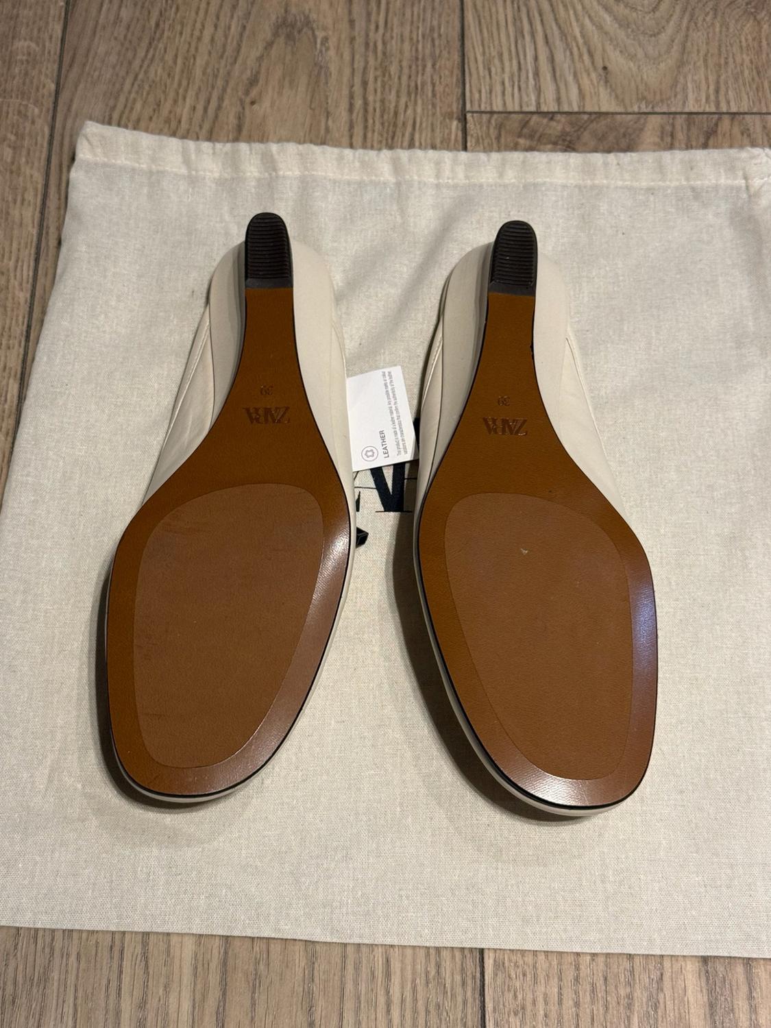 Pair of white shoes viewed by their brown wedge soles, resting on a fabric bag on a wooden floor. Text: ZARA, 39, LEATHER.