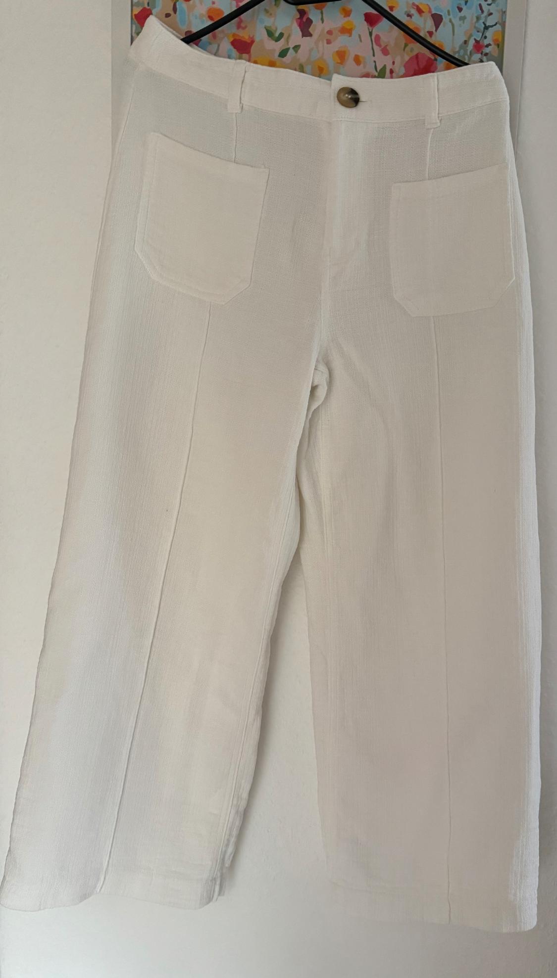 White lightweight fabric trousers with front pockets, hanging on a hanger against a colourful floral background.