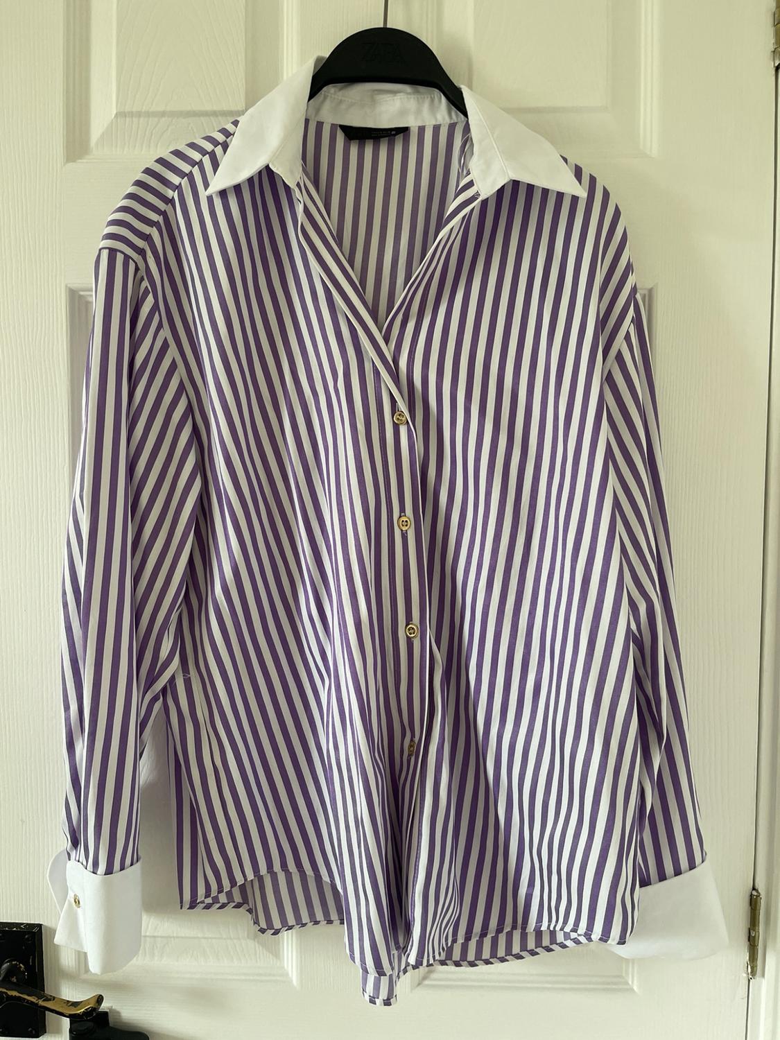 Purple and white striped shirt, white collar and cuffs, and gold buttons. It hangs from a black hanger that says ZARA on a white door.
