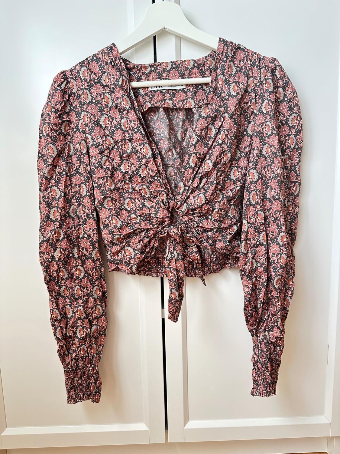 Pink floral print long-sleeved crop top. V-neck, front knot, and elasticated cuffs. Hanging on a hanger in a white wardrobe.