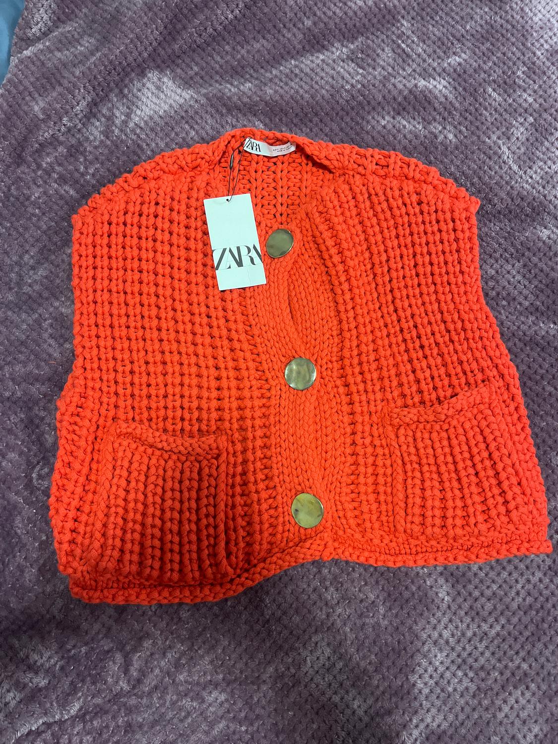 Orange knitted waistcoat with gold buttons and front pockets over purple fabric. Label with "ZARA" text hanging.