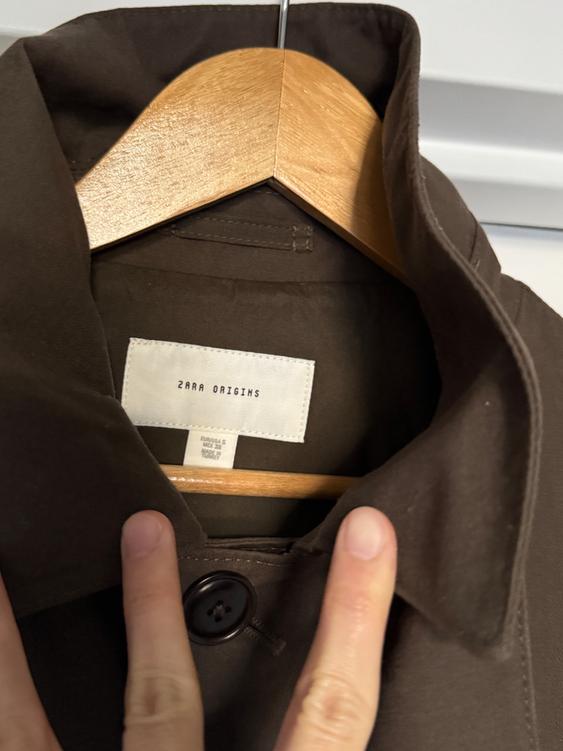 Brown coat on a wooden hanger with a "ZARA ORIGINS" tag. Two fingers hold the collar, showing a dark button.