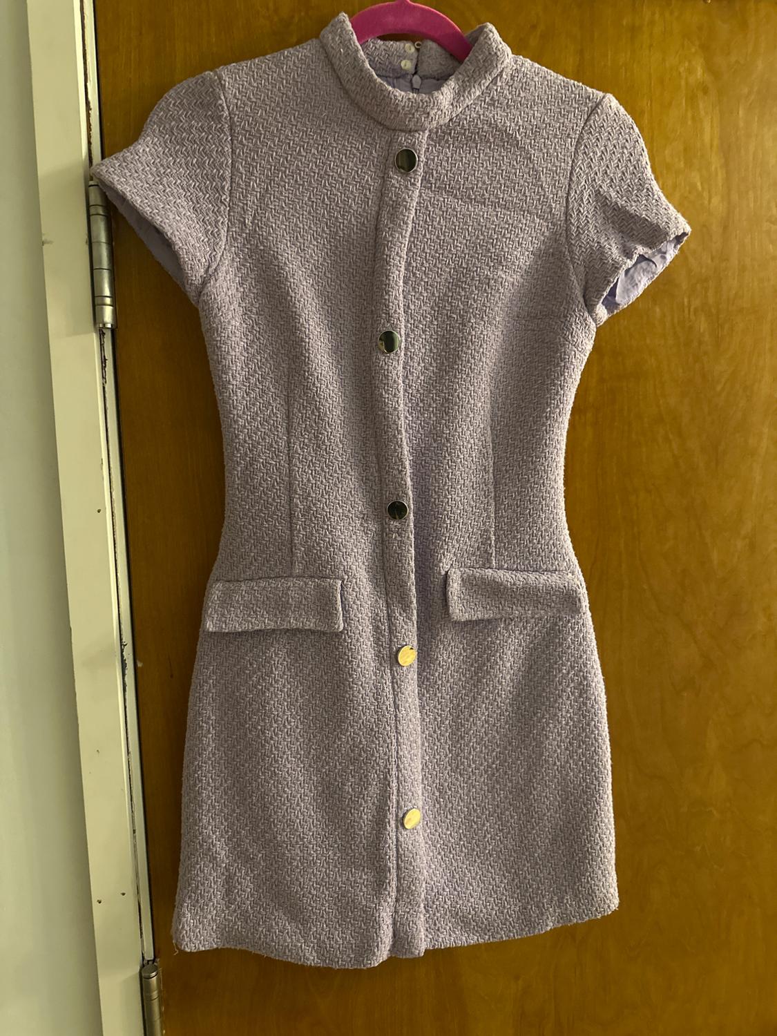 Textured lilac short-sleeved dress with a high neck, buttons and front pockets. It hangs from a pink hanger on a wooden door.