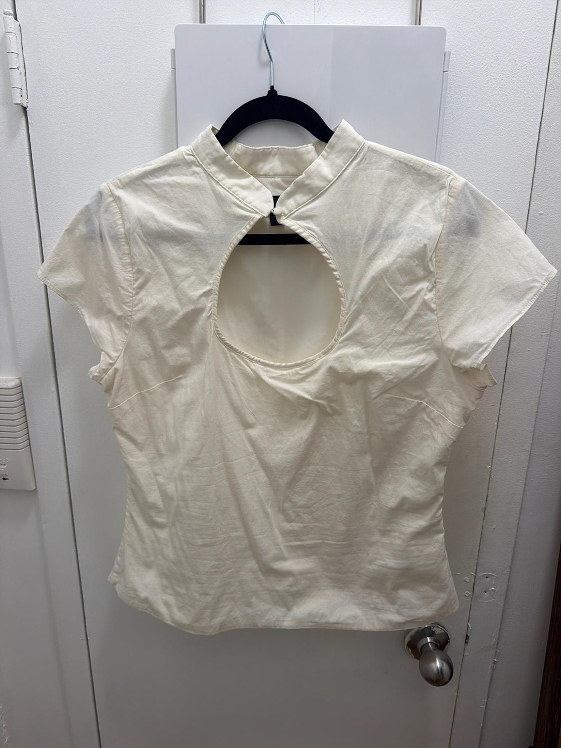 Cream-colored short-sleeve blouse with a high neck and circular opening at the chest, hanging on a black hanger over a white door.