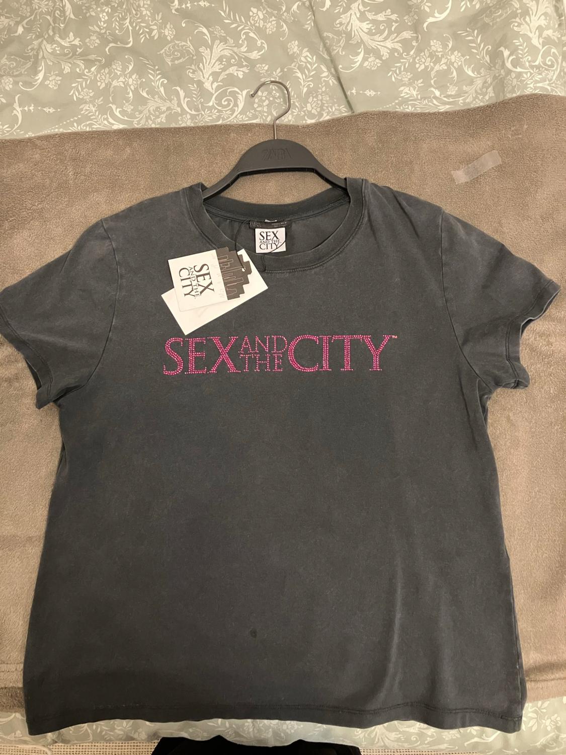 Dark grey t-shirt on a bed. The text "SEX AND THE CITY" stands out in pink. Includes a "SEX AND THE CITY" label and a "Zara" hanger.