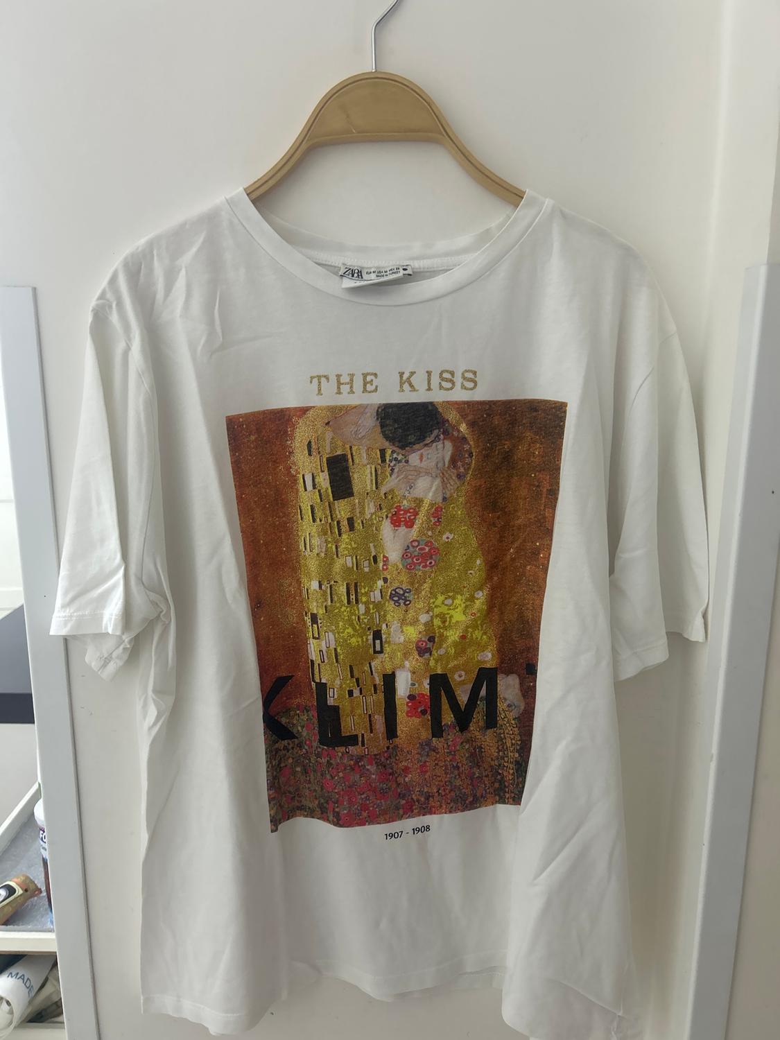 White hanging T-shirt with a print of Klimt's "The Kiss" on the front. Visible text: "THE KISS", "KLIMT", "1907-1908".