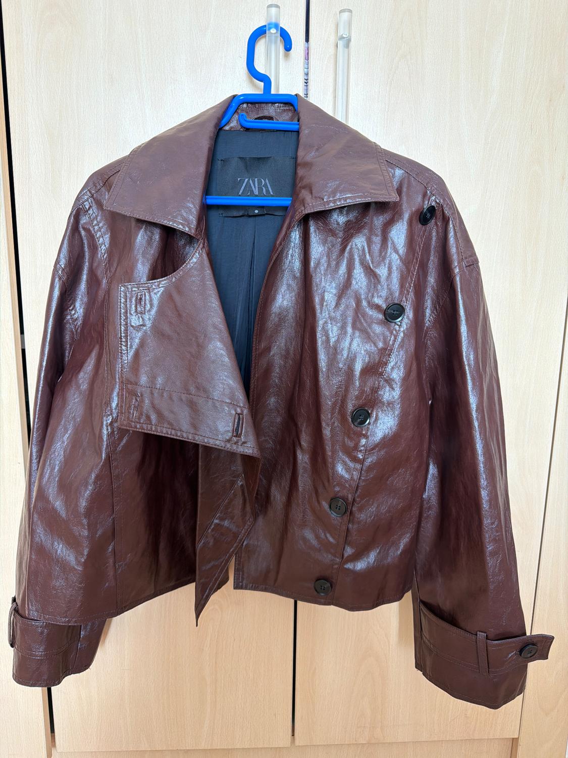Brown faux leather jacket with black buttons, hanging on a blue hanger. Visible inner label with the text "ZARA".