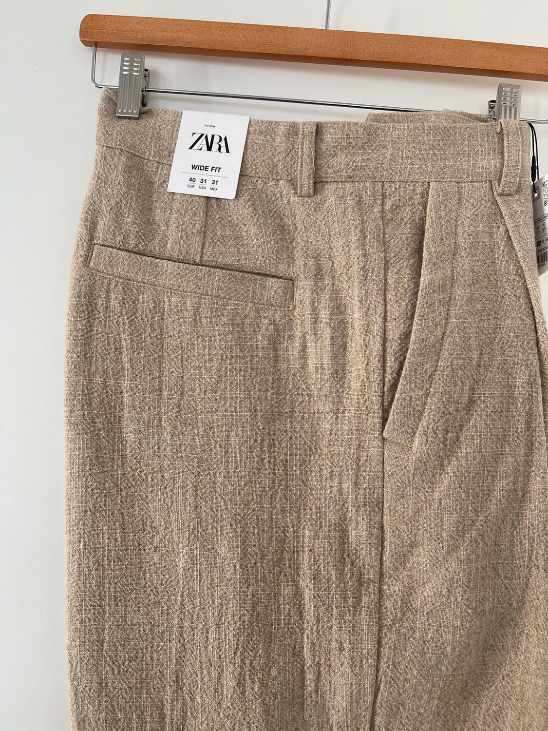 Close-up of textured beige trousers on a wooden hanger. Label with the text: ZARA WIDE FIT 40 31 31 EUR USA MEX.