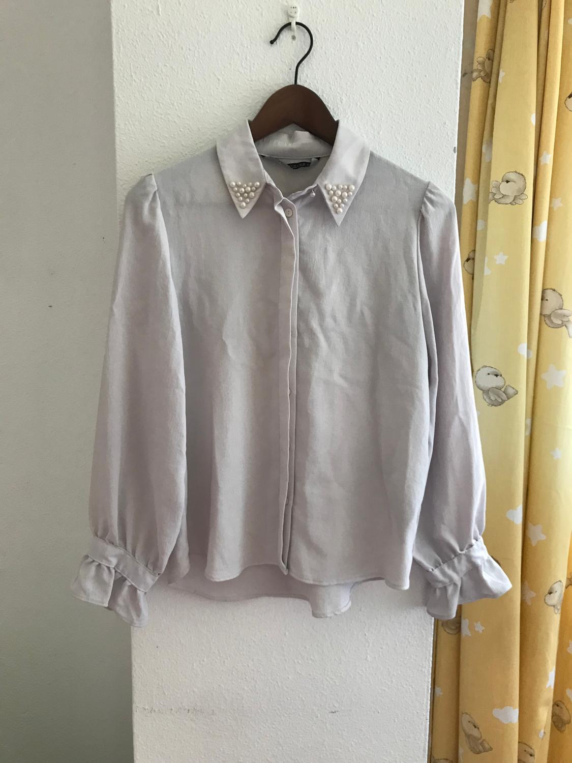 White blouse hanging on a wooden hanger, with a pearl-decorated collar. White wall and yellow patterned curtain in the background.