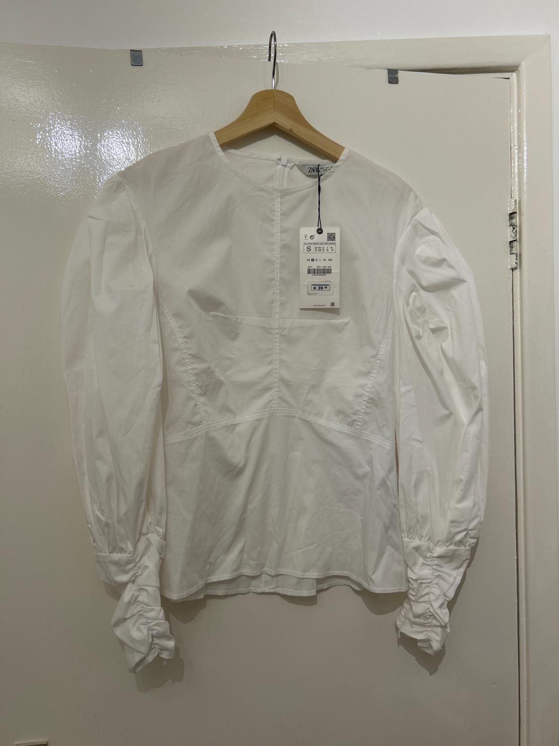 White long-sleeved blouse with a hangtag, hanging on a wooden hanger over a white door.