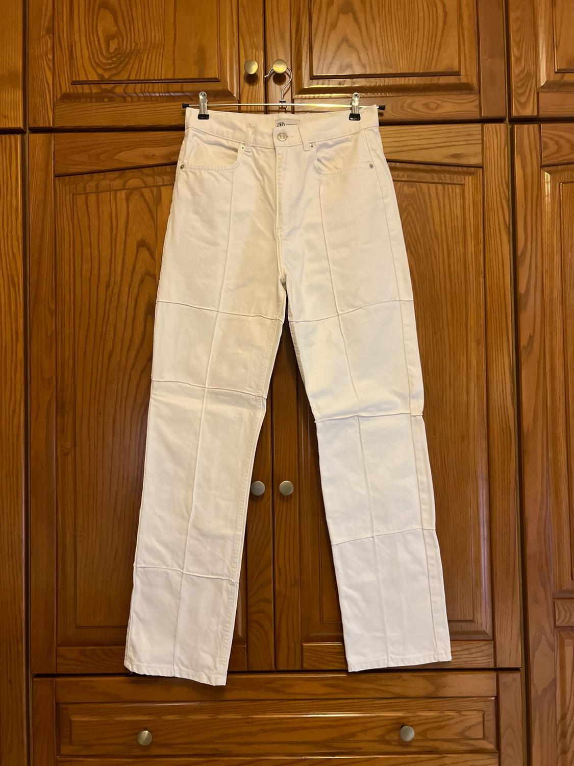 White trousers with panelled seams hanging in front of a wooden wardrobe. ZARA label visible. Casual and homely style.