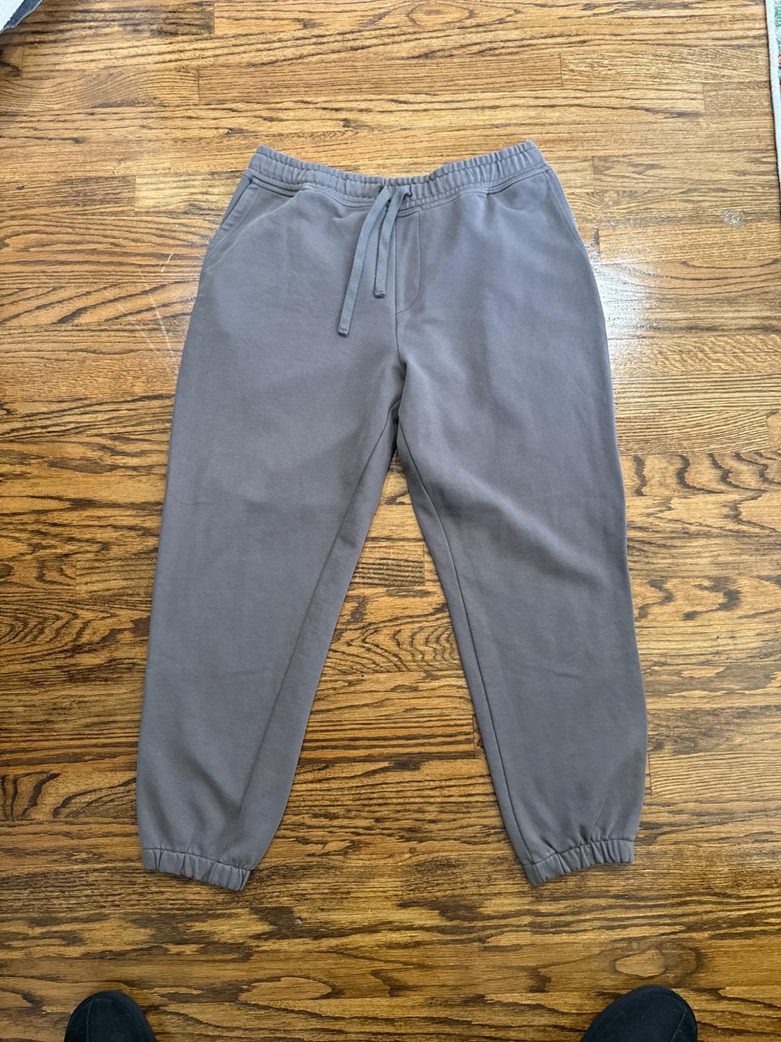 Gray track pants with a drawstring and elastic cuffs, on a wooden floor. Two black shoes peek out from the bottom edge.