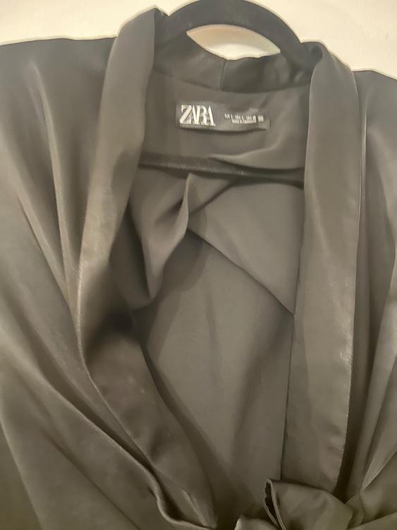Close-up of a black garment hanging on a hanger, with a visible label that reads "ZARA". Soft, shiny fabric.