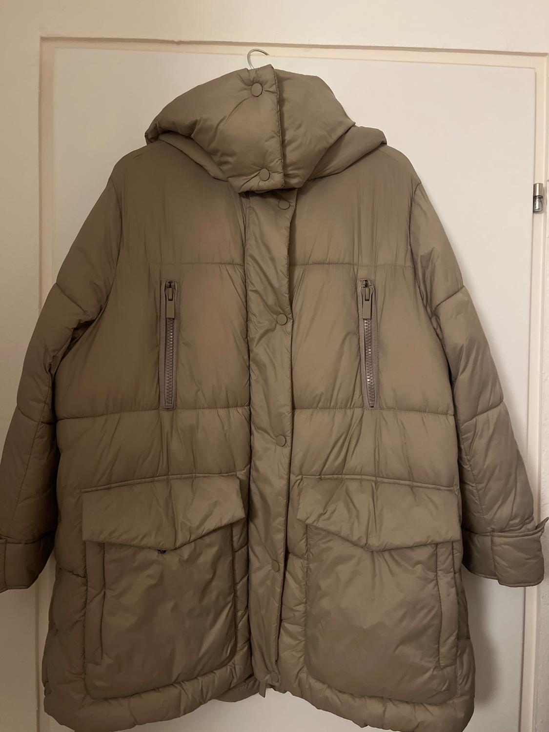 Beige padded coat with a hood, button fastening, and zips on the pockets, hanging on a white door.