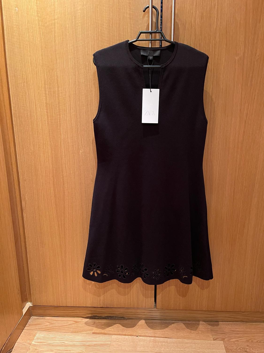 Black sleeveless dress hanging on a wooden wardrobe. It features a hem with die-cut flowers and a label with the text ZARA.