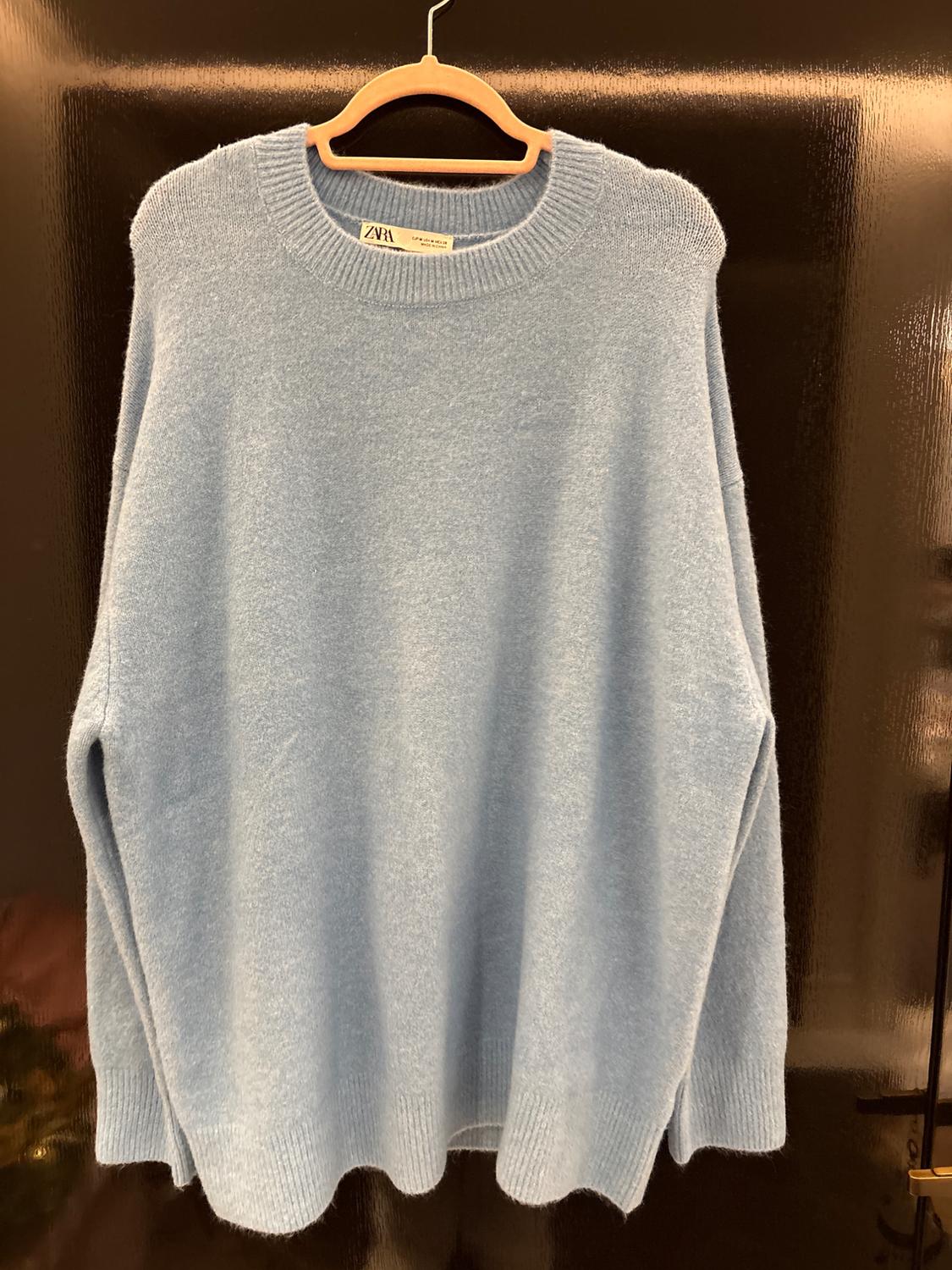 Light blue jumper hanging on a wooden hanger against a dark background. Visible label with the text "Zara".