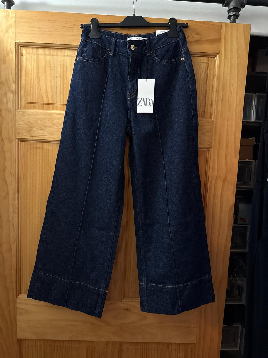Dark blue wide-leg jeans with a front seam and a ZARA label, hanging on a hanger in front of a wooden door.