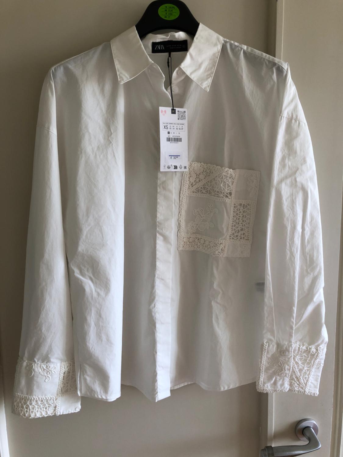 White, long-sleeved shirt with embroidered detailing on the pocket and cuffs, hanging on a black hanger with a visible Zara label.