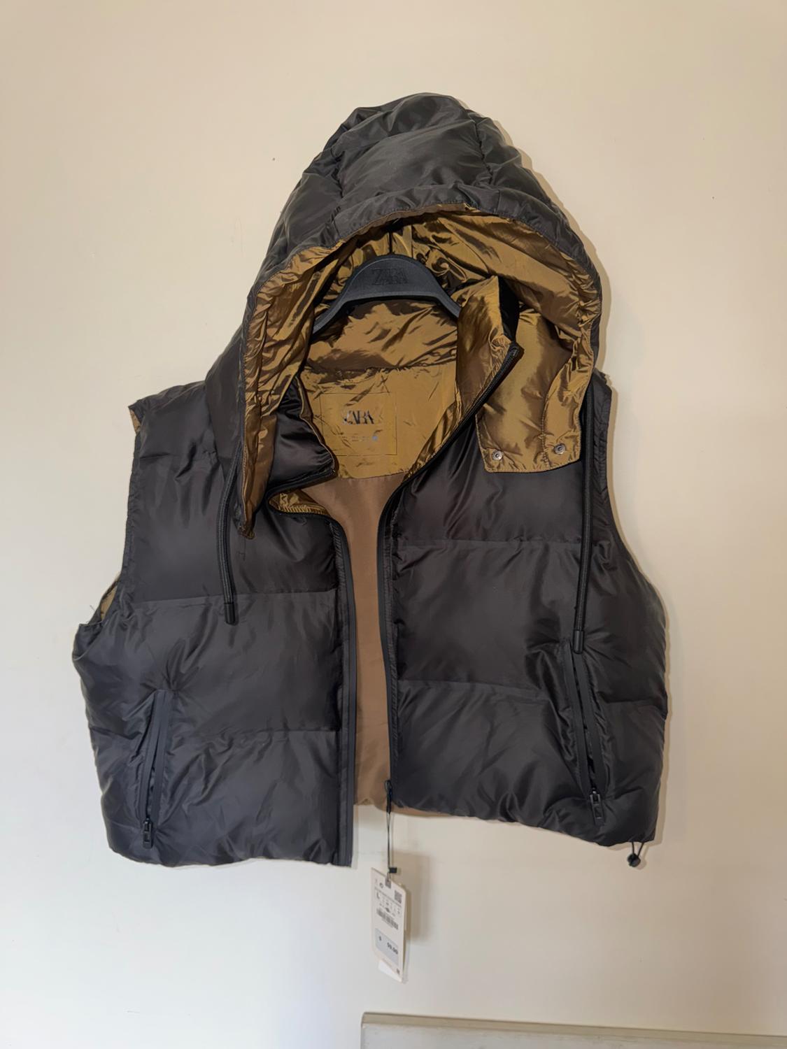 Dark grey padded gilet with a hood and gold-coloured lining hanging on a light-coloured wall. ZARA text on the label and hanger. Casual style.