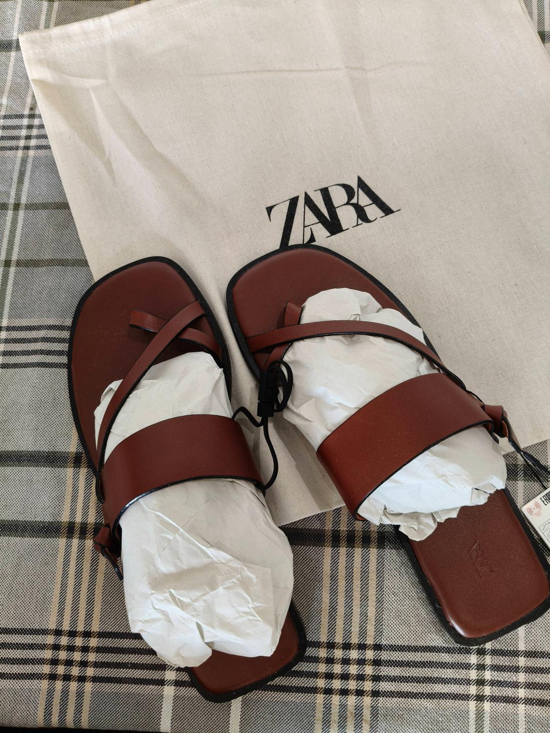Brown flat strappy sandals stuffed with paper, on a white bag bearing the text ZARA, against a checked fabric background.