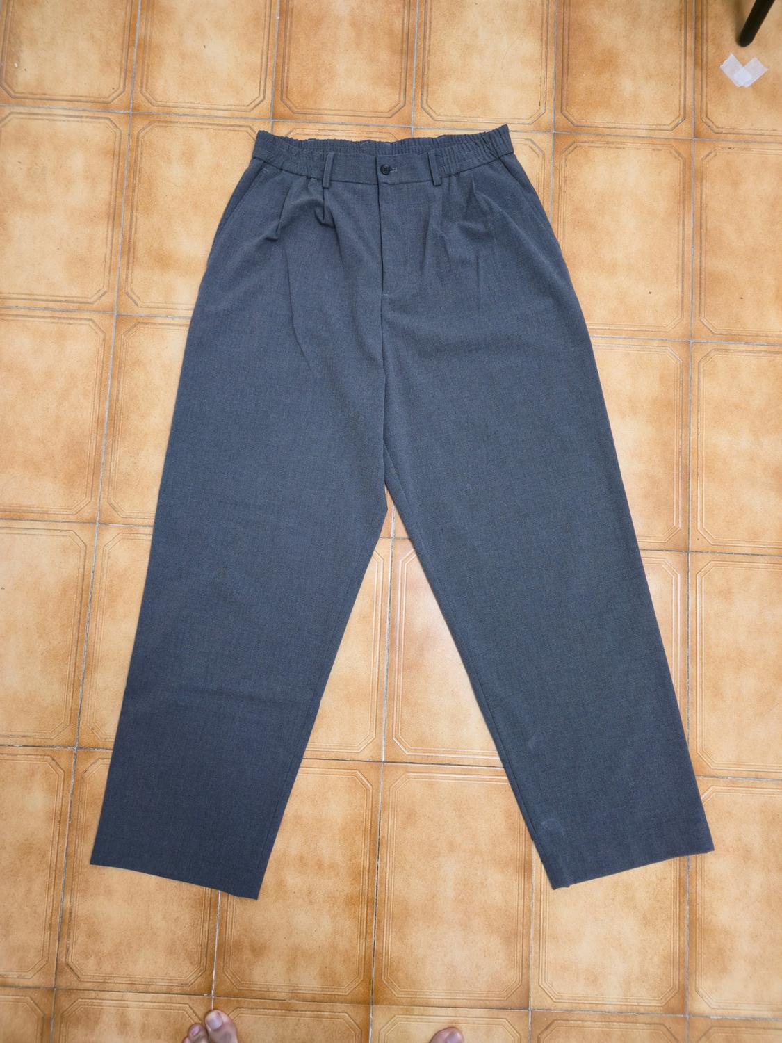 Grey, straight-cut trousers with an elasticated waist and pleats, laid on a brown tiled floor.