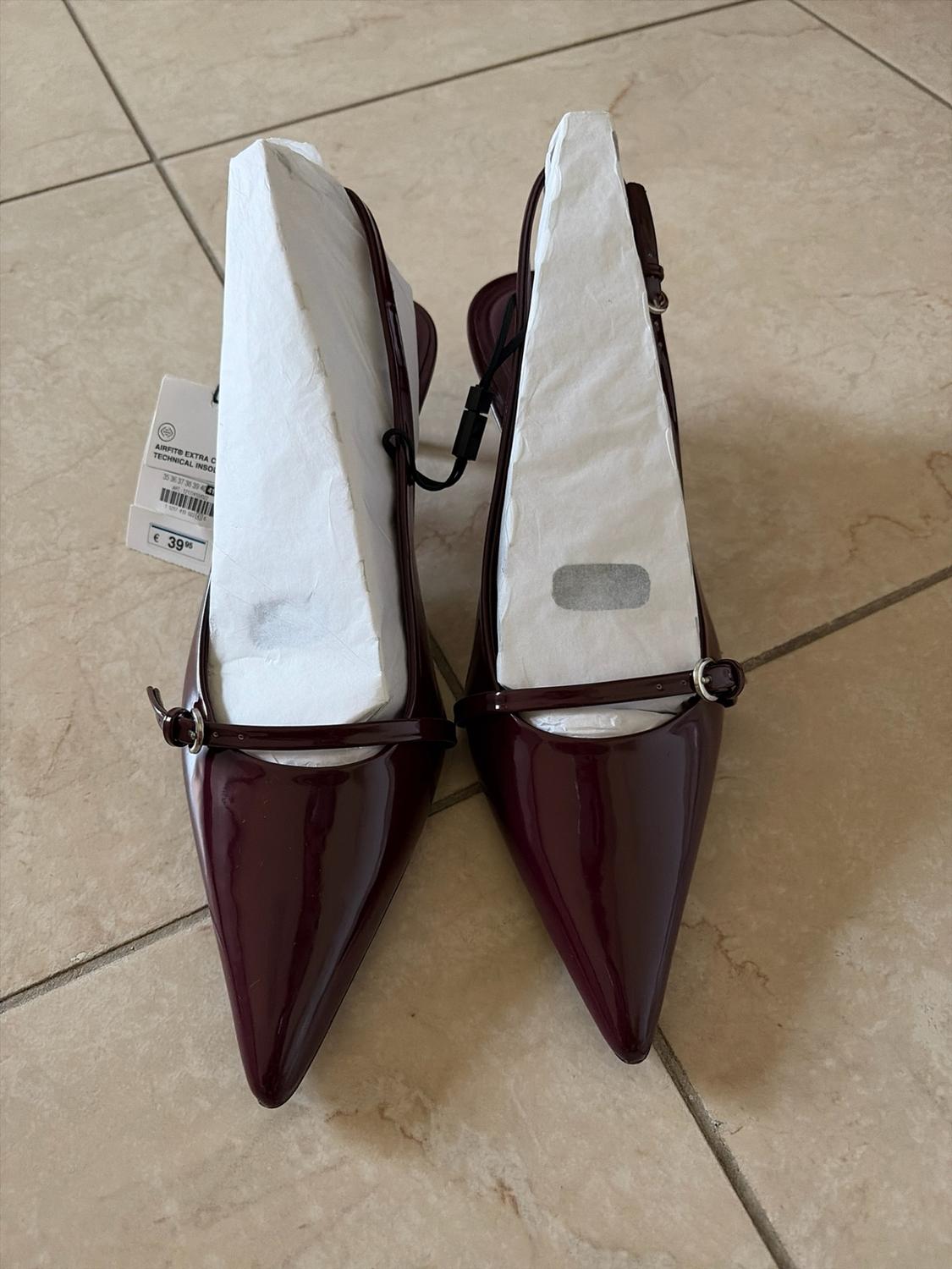 Burgundy patent stiletto heels with a pointed toe, an instep strap and paper stuffing on a tiled floor. Label: € 39.95.