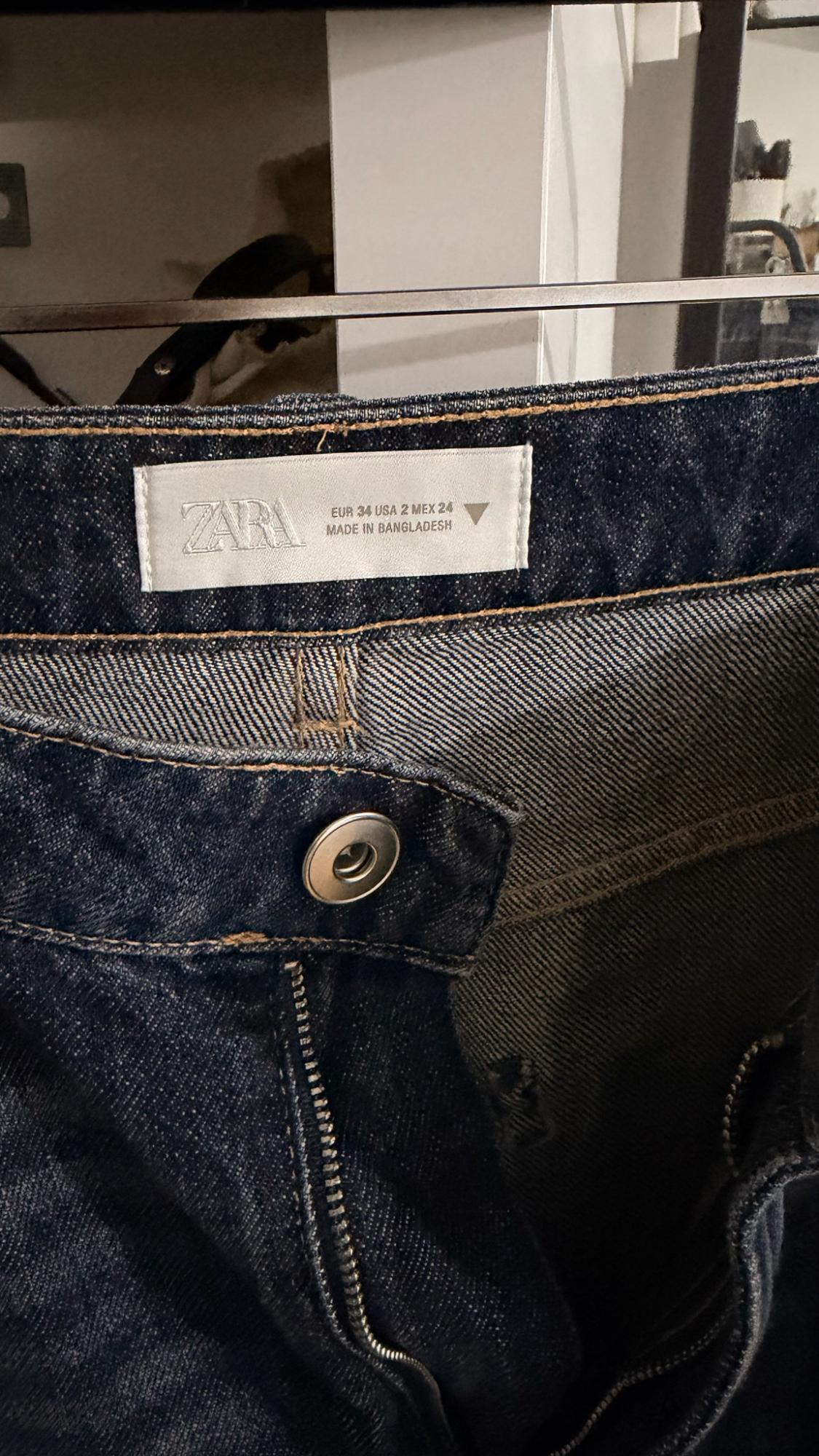 Blue jeans hanging. Label with text: ZARA EUR 34 USA 2 MEX 24 MADE IN BANGLADESH. Detail of silver button and zip.