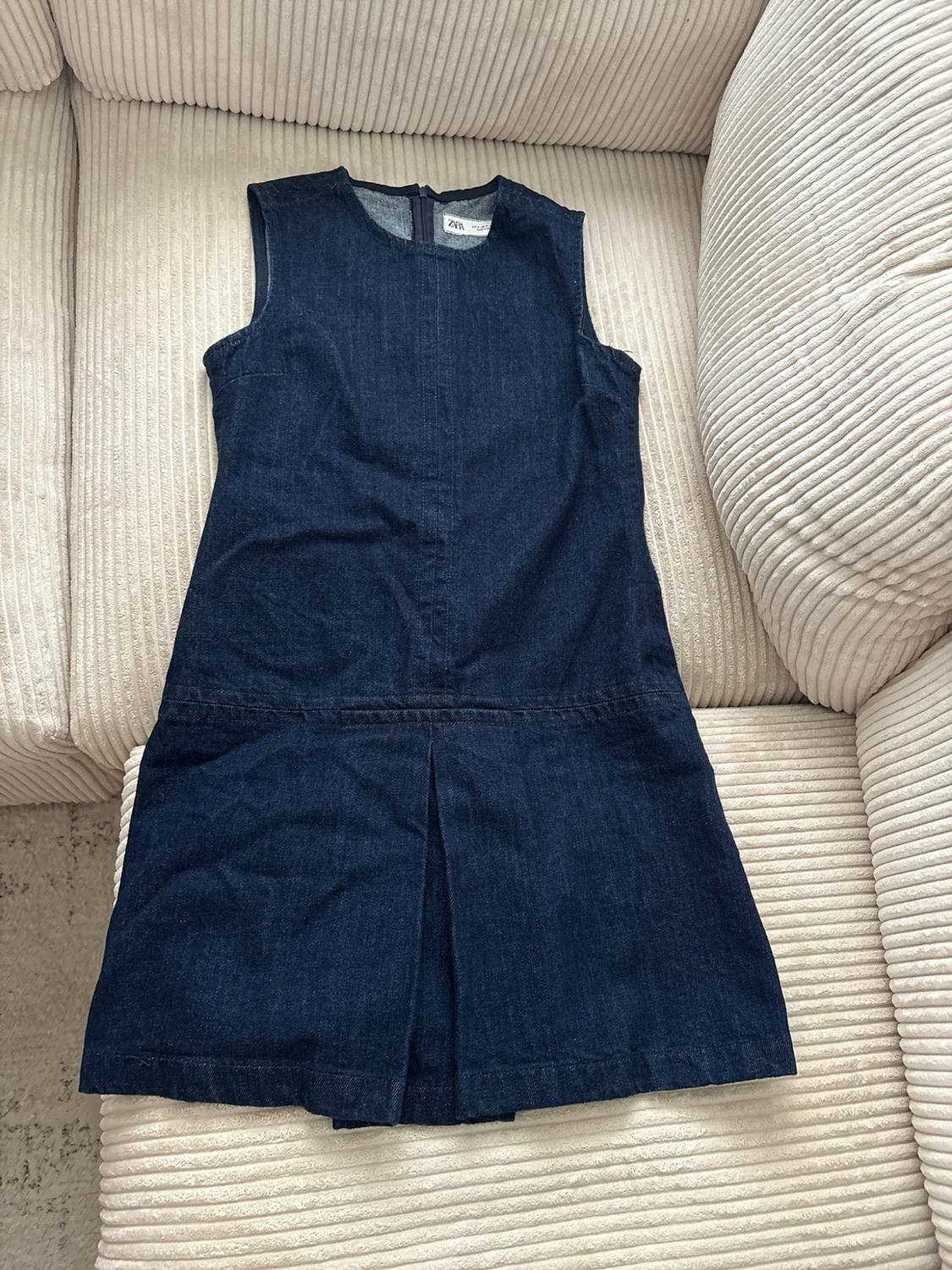Dark blue sleeveless denim dress with a round neck and pleated hem. Visible ZARA label. Resting on a beige corduroy sofa.