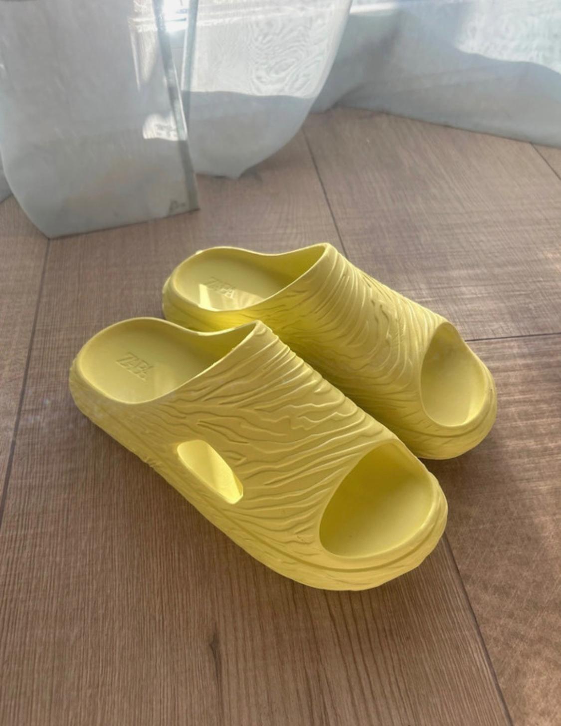 Yellow sandals with a wavy texture and ZARA text on the insole, on a wooden floor with natural light and a translucent curtain in the background.