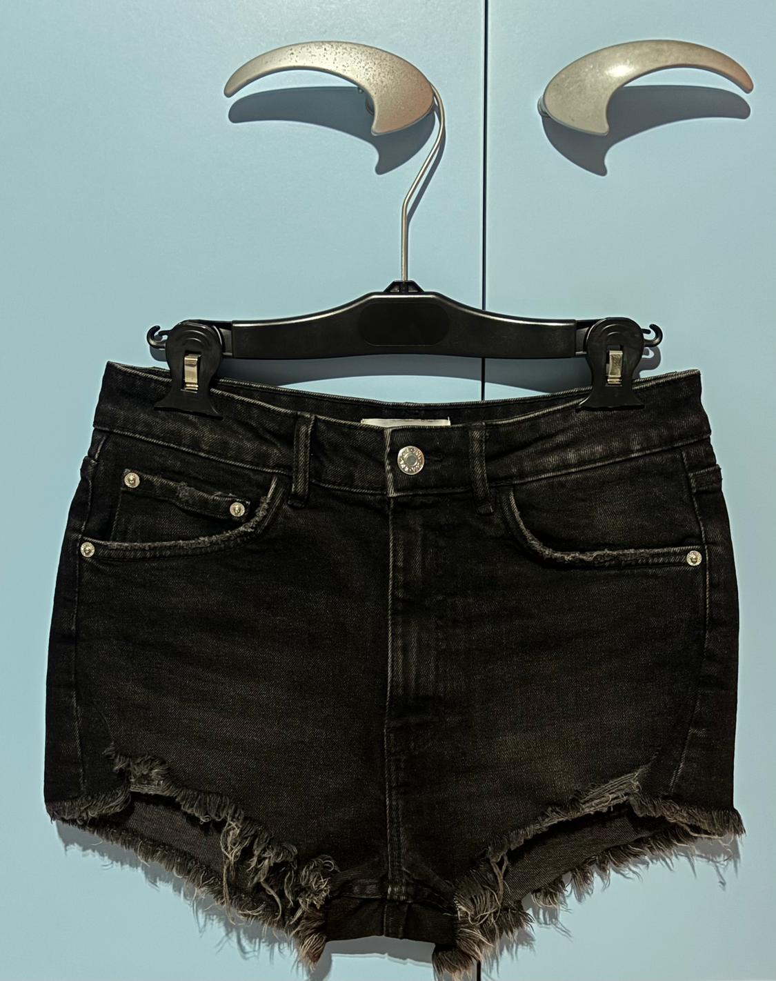 Μαύρο σορτς από denim με ξεφτισμένα άκρα, κρεμασμένο σε μια μαύρη κρεμάστρα, πάνω σε ένα γαλάζιο φόντο.