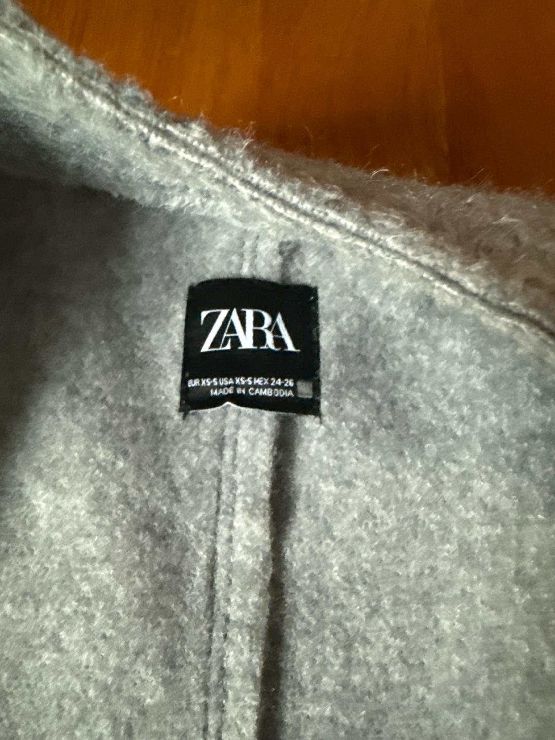 Gray clothing tag with the text "ZARA EUR XS USA XS MEX 24-26 MADE IN CAMBODIA" on a black background. Soft textured fabric.