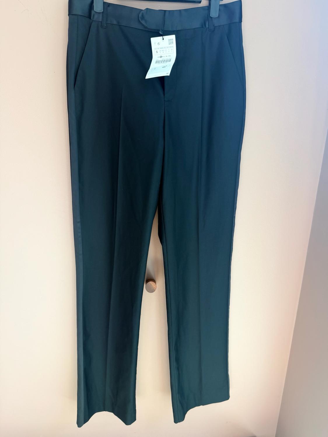 Dark trousers with a satin waistband and side stripe, hanging on a black hanger in front of a light-coloured wall. White label visible.
