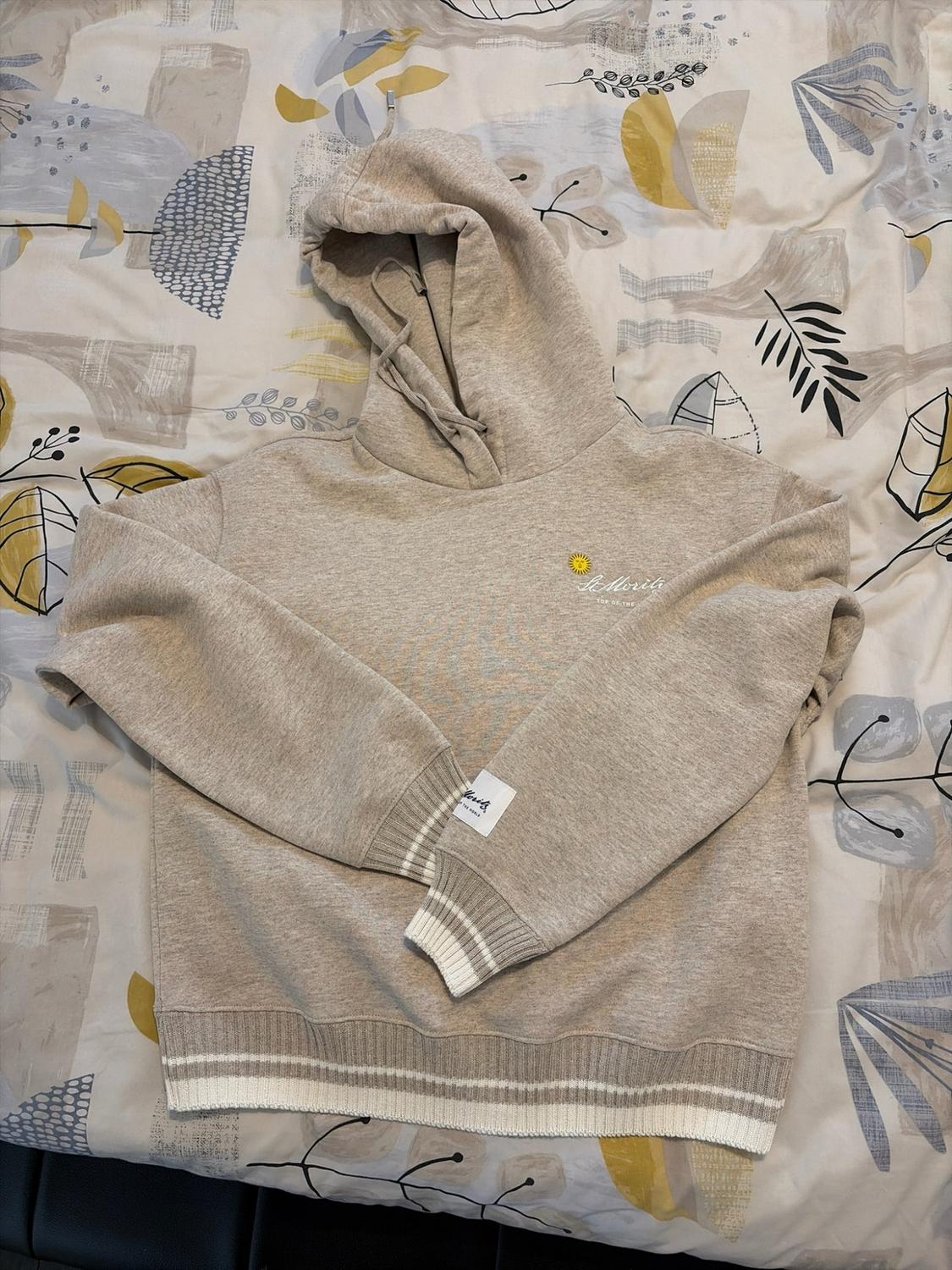 Beige hooded sweatshirt with striped cuffs and hem. "St. Moritz TOP OF THE WORLD"
embroidery and a sun on the chest, over a printed patch.