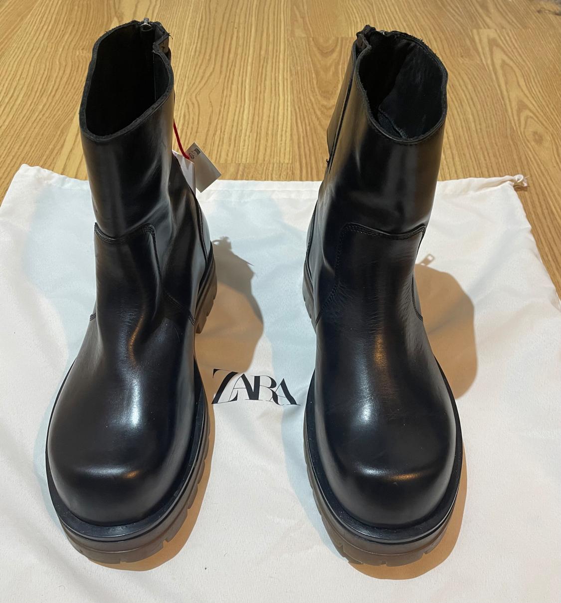 Black leather boots on a white bag with the "ZARA" logo, placed on a light wooden floor.
