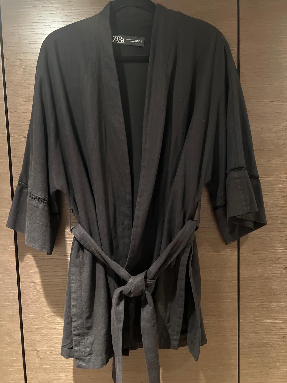 Black, short-sleeved kimono with a knotted belt, hanging on a hanger in front of a wooden background. Visible label: Zara.
