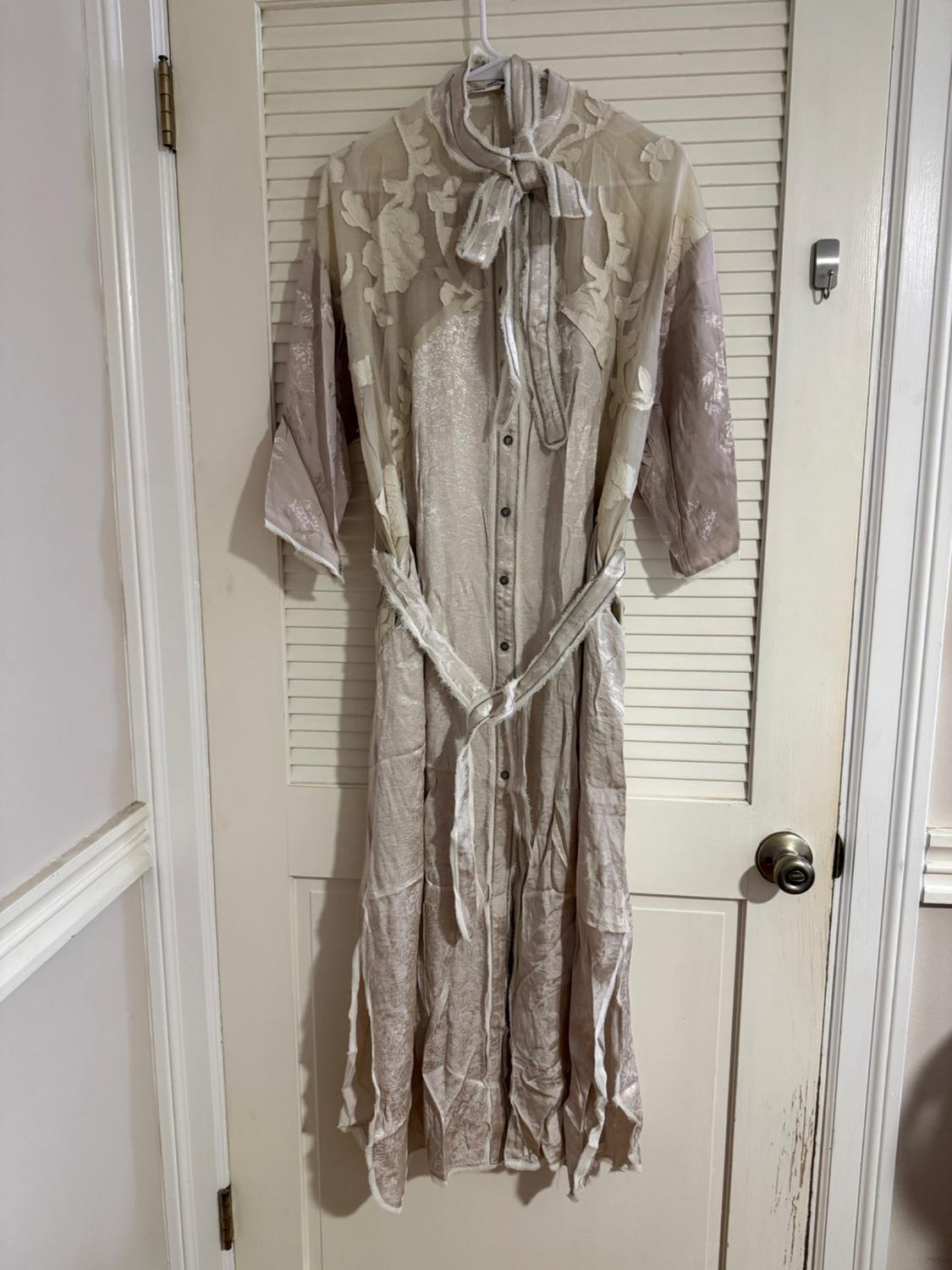 Beige dress with a floral print and bow at the neck, hanging on a white paneled door. Three-quarter sleeves and front buttons.