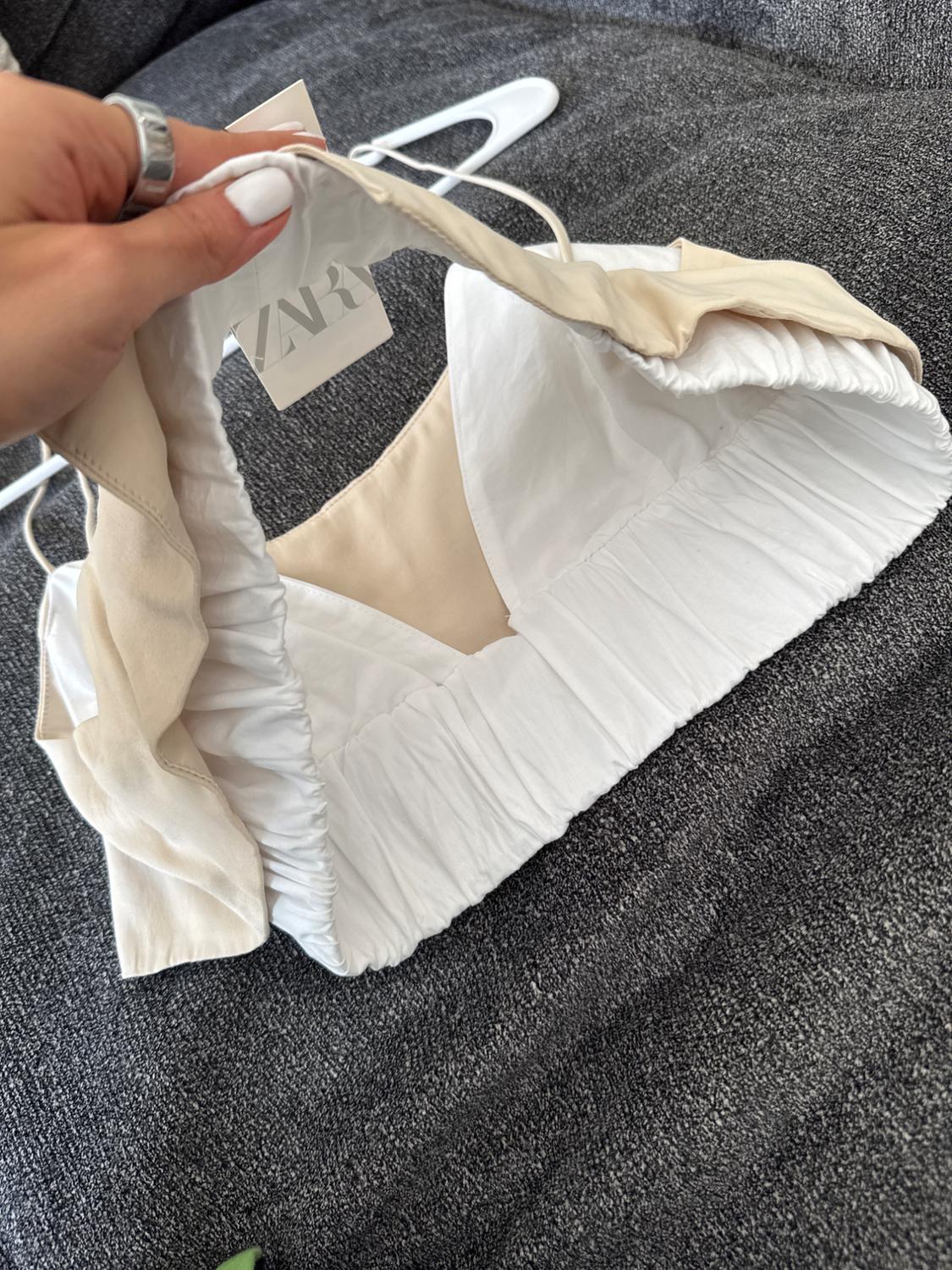 Hand holding a white and beige top with visible ZARA tag, on a gray fabric background.