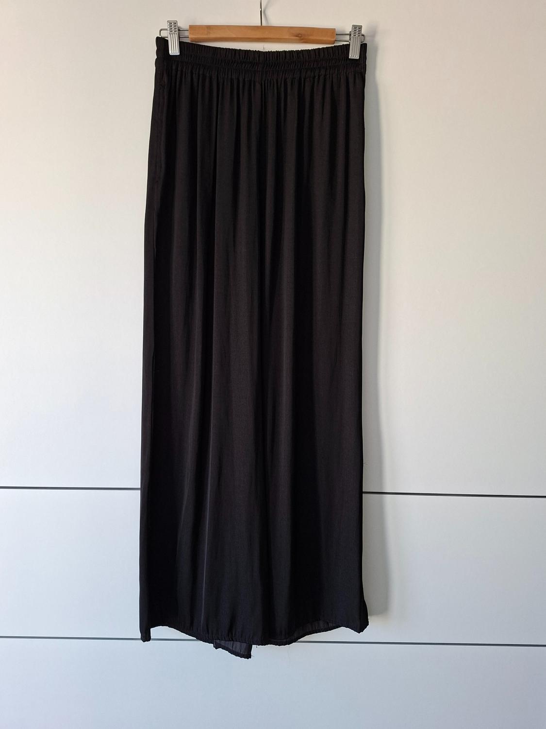 Flowing black wide-leg trousers hanging from a wooden hanger against a white background with horizontal lines.