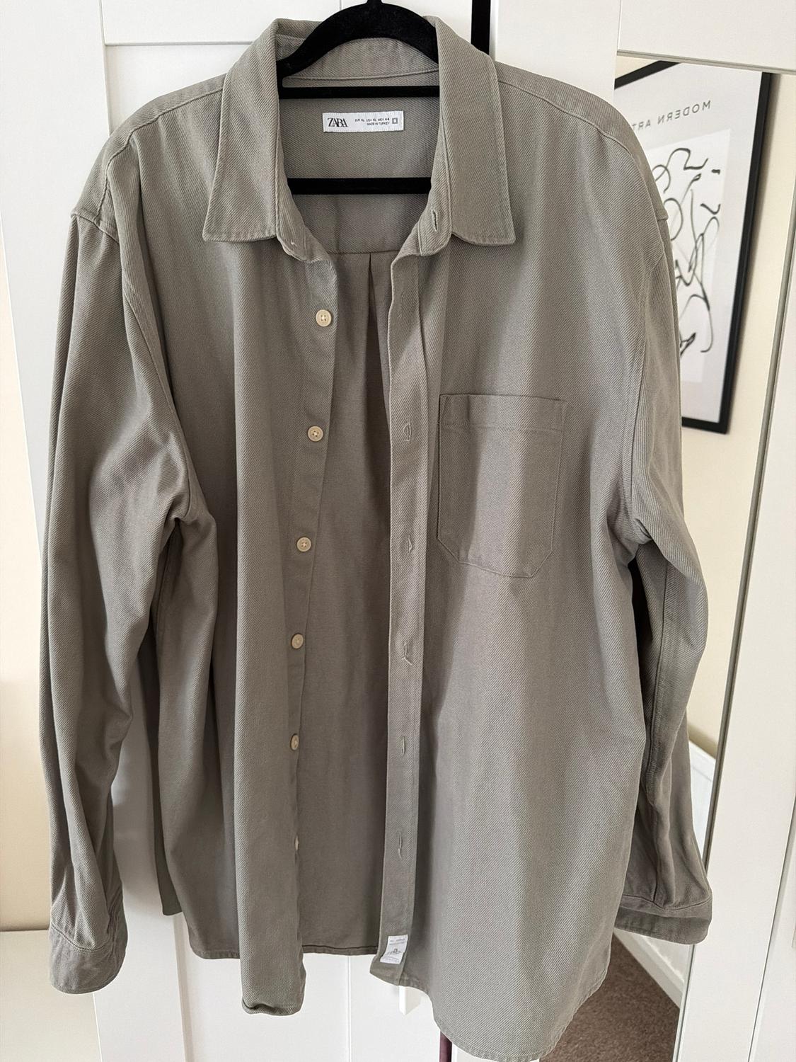 Grey long-sleeved shirt hanging on a black hanger. Background with a white door and a modern art picture on the wall.