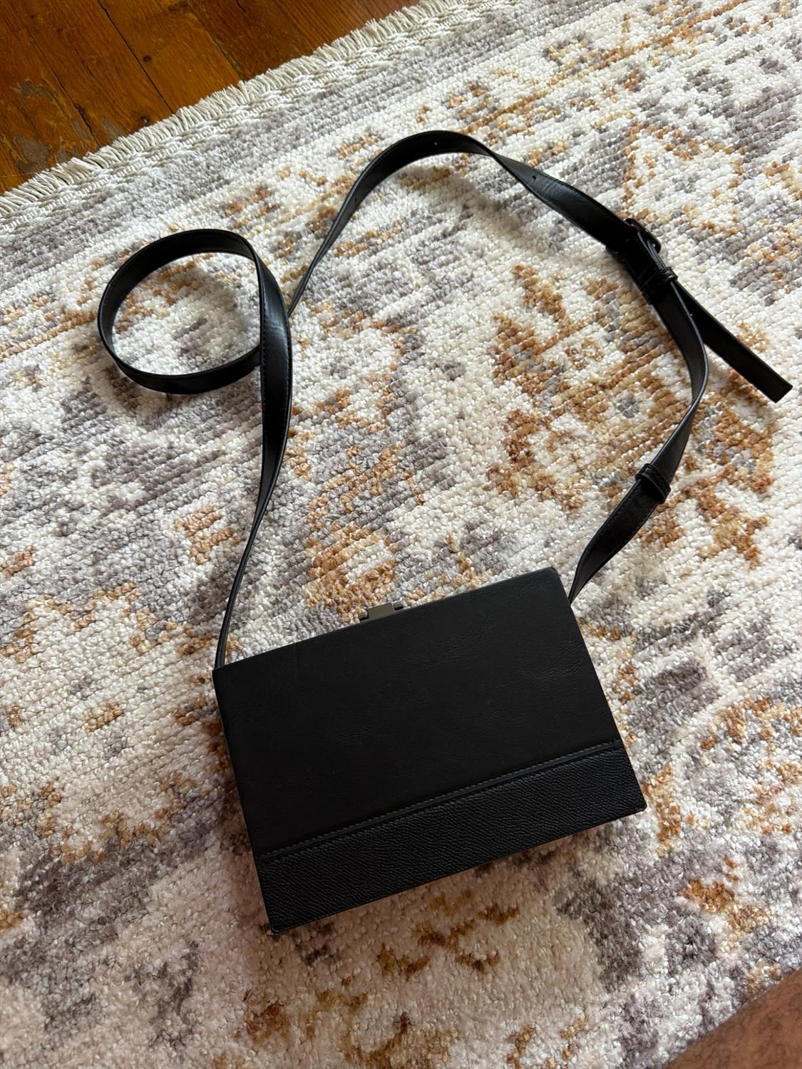 Black rectangular handbag with a long strap and mixed texture, resting on a neutral-toned patterned rug.