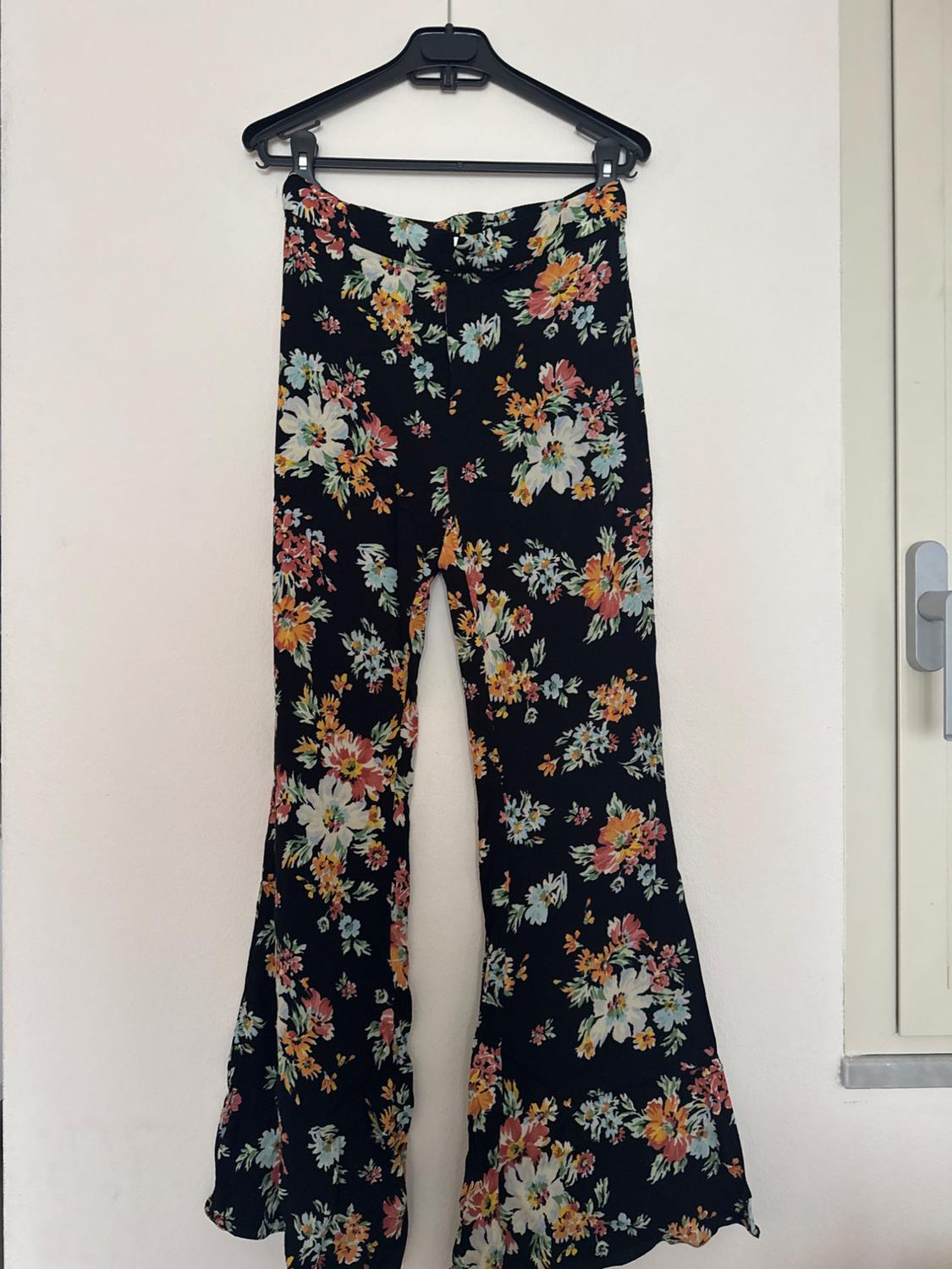Black fabric flared trousers with a colourful floral print, hanging on a hanger against a white wall.