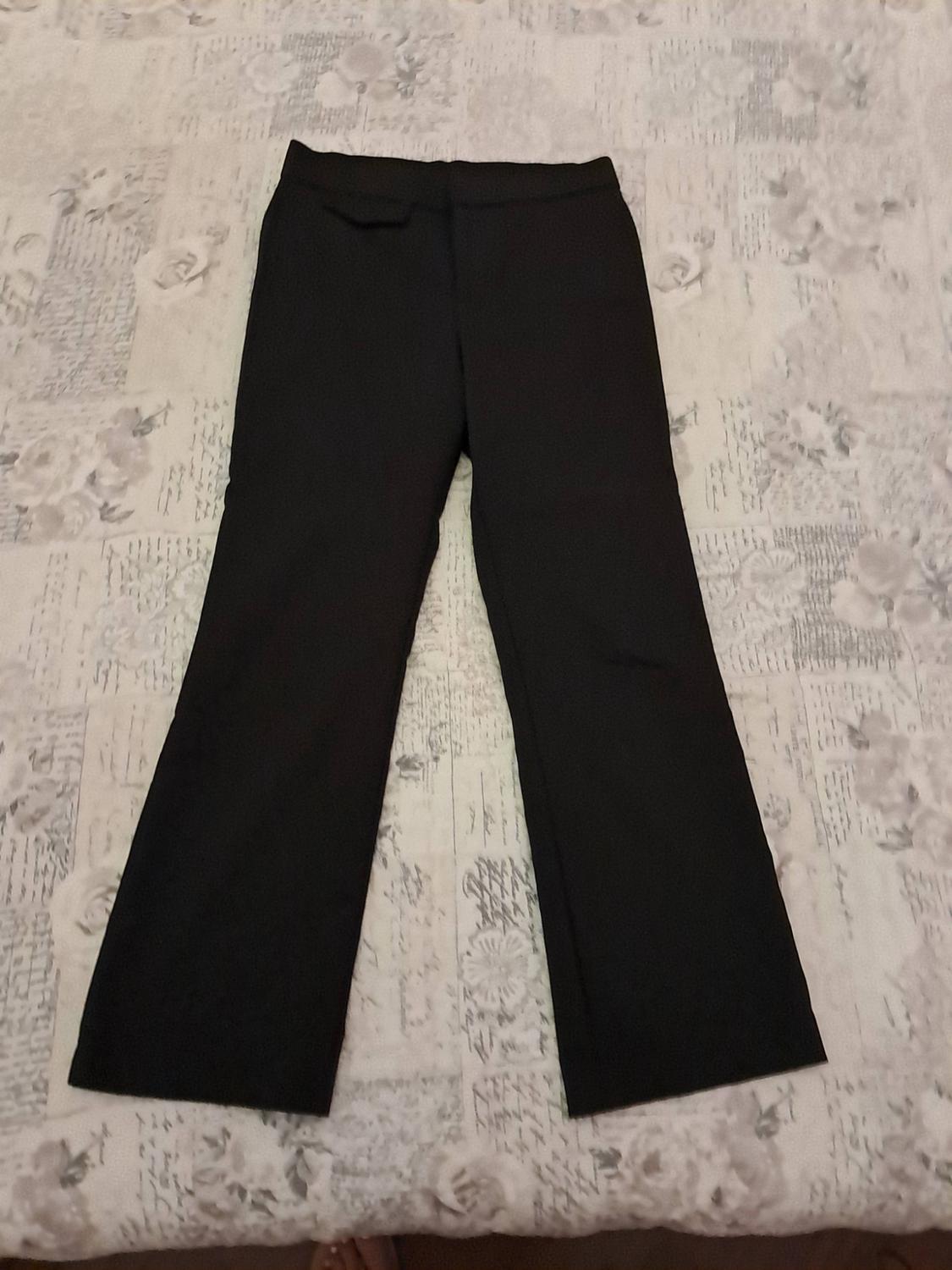 Long black trousers laid out on a surface with a light-coloured pattern. They feature a flap pocket detail at the waistband.