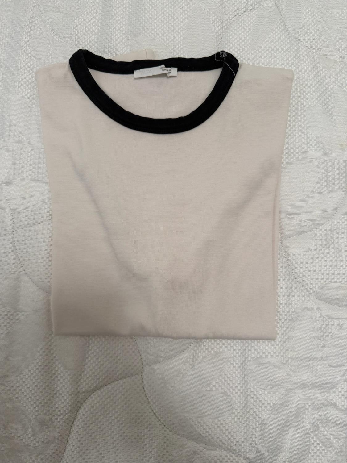 Βασικό ριχτό t-shirt σε εκρού χρώμα με μαύρο τελείωμα στη στρογγυλή λαιμόκοψη, σε λευκή, υφασμένη επιφάνεια.